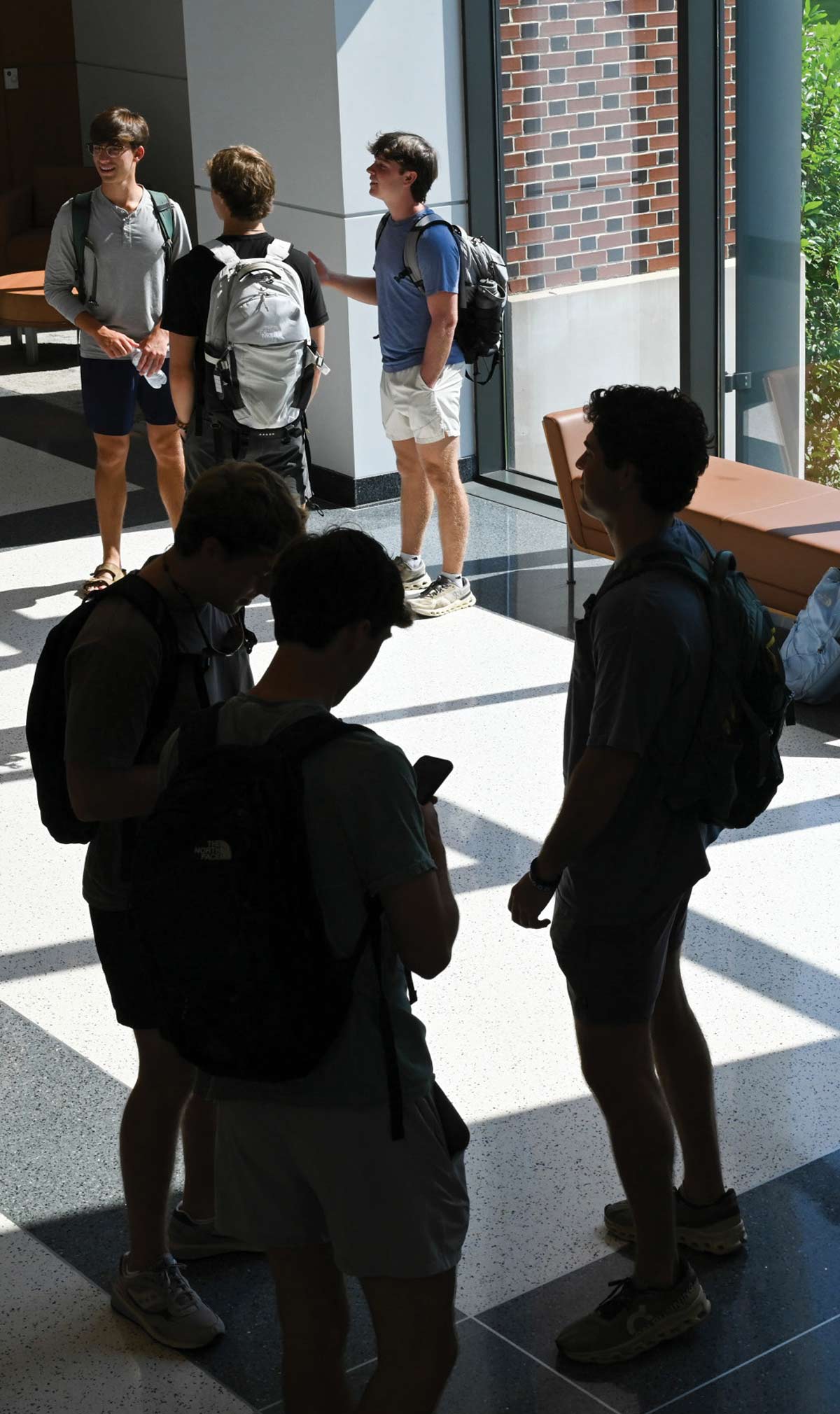 Indoor shot of a group of male college students in a modern, sunlit university hallway lobby area; All of them are dressed casually with backpacks