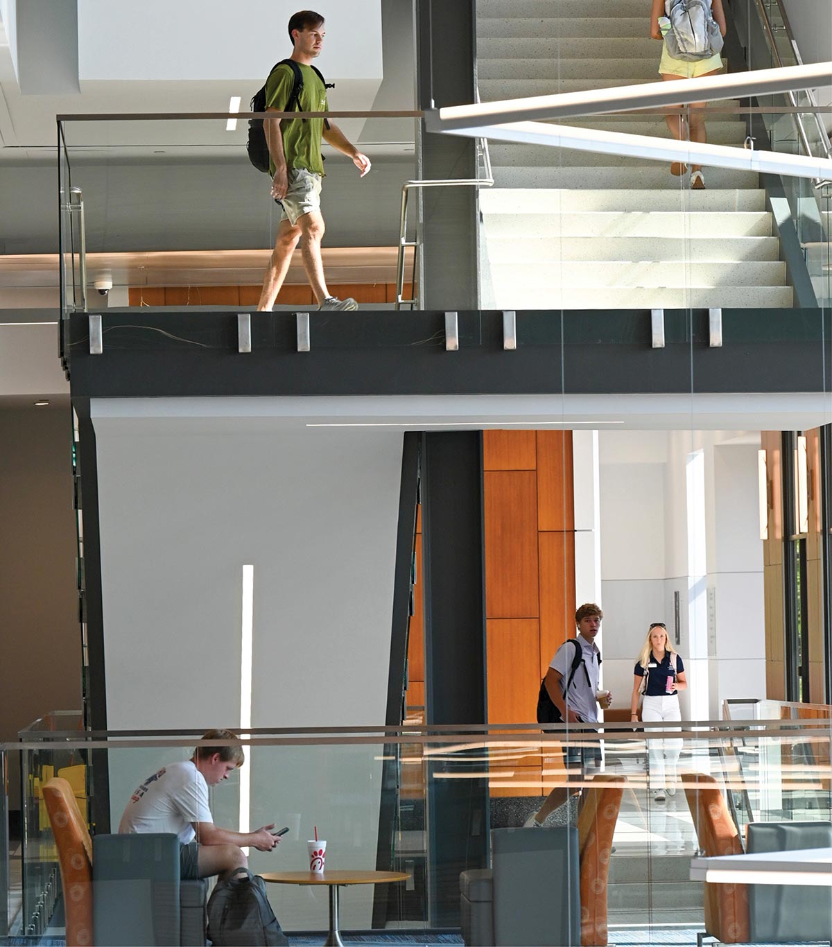 A vertical, multi-level indoor shot of a modern university building showing a few college students walking and another student looking down at his phone seated down in a chair