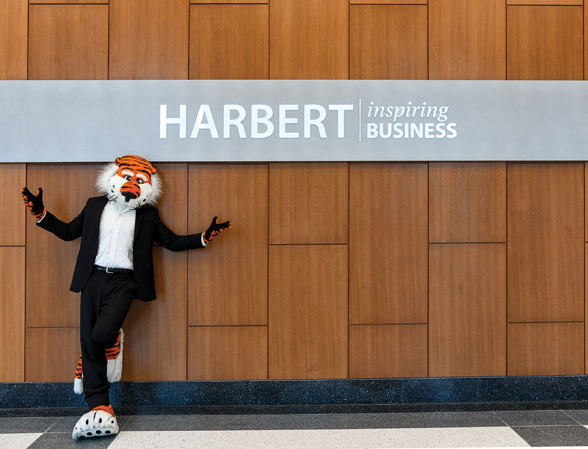 Auburn University's tiger mascot in a business suit attire leaning against a wood-paneled wall and above the mascot is a sign that reads HARBERT | inspiring BUSINESS