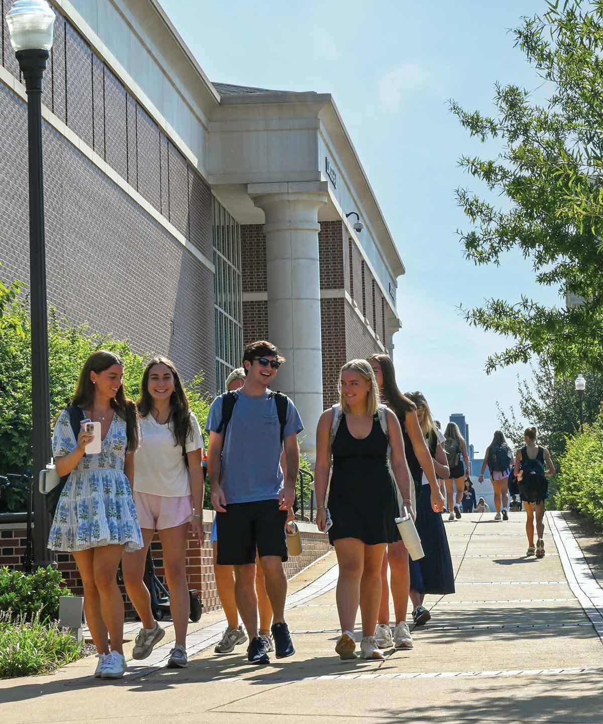 A diverse group of college students walking along a paved campus walkway on a bright, sunny day as they are situated nearby a large brick university building with white columns
