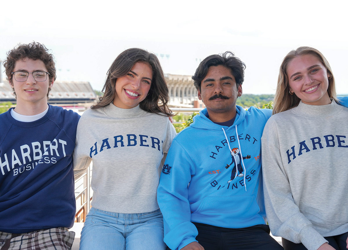 Four students sitting side-by-side and smiling in different various Auburn Harbert College of Business merchandise