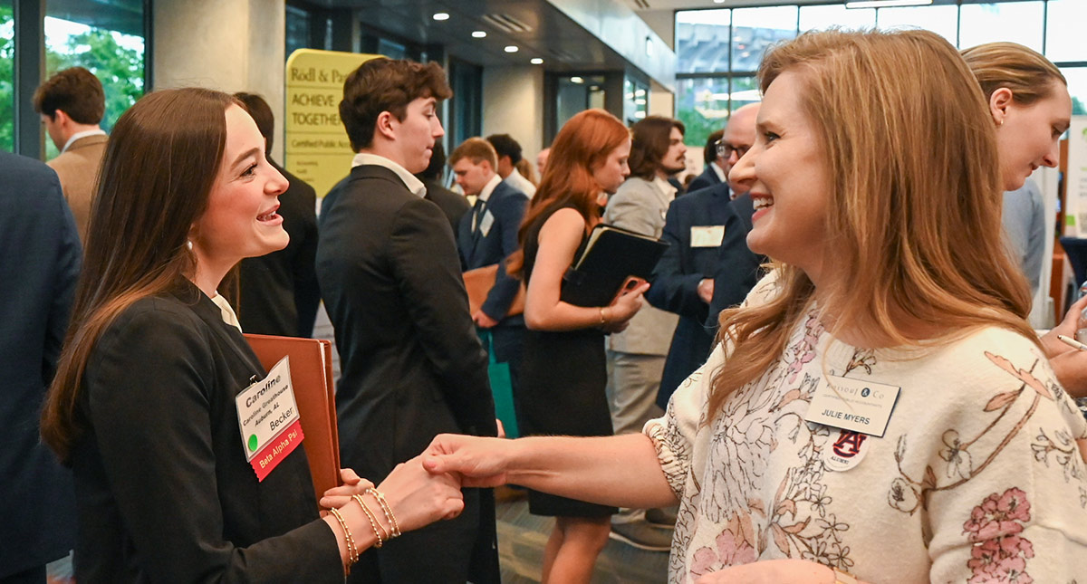 Two women in professional attire shake hands and smile at a career fair. Both wear name tags. Other students and recruiters in suits fill the background of the hall.