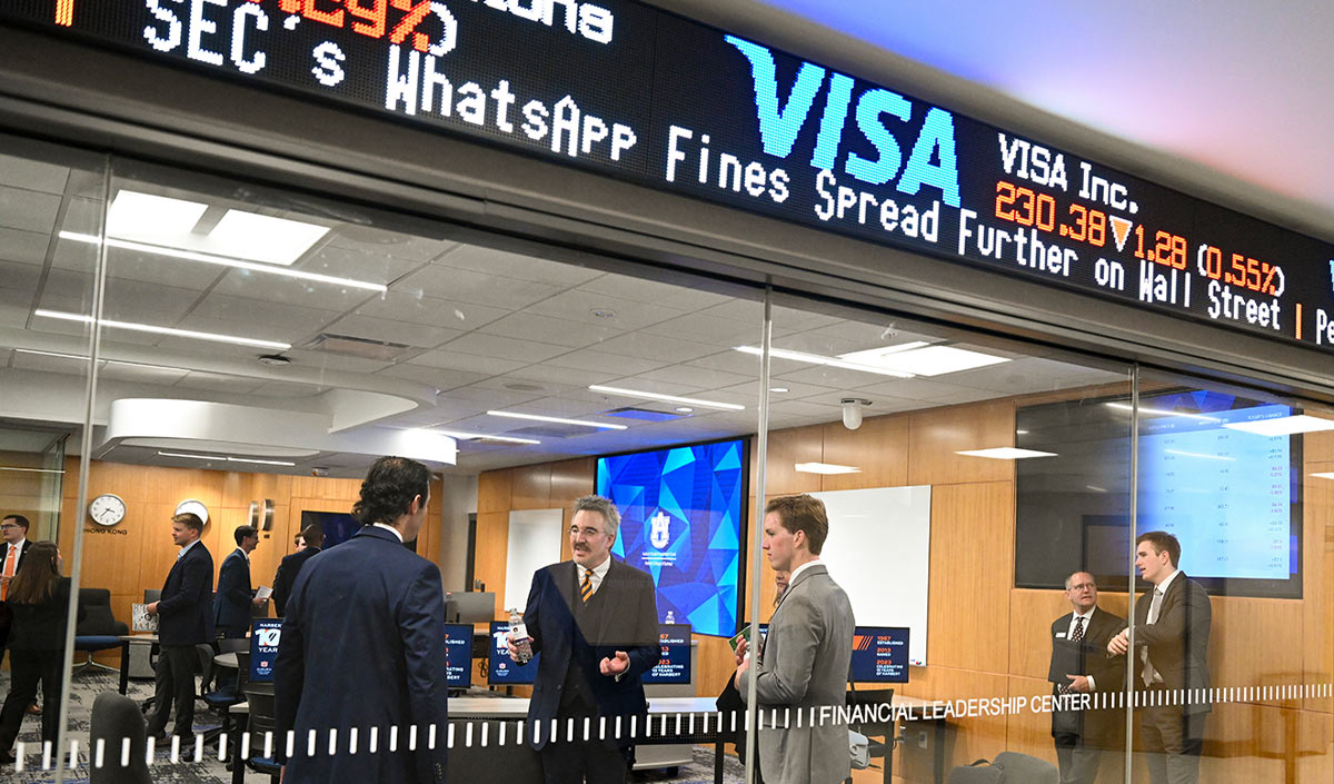 Professionals and students converse inside the glass-walled Financial Leadership Center. A large electronic ticker above the room displays live stock market data for Visa and other firms.