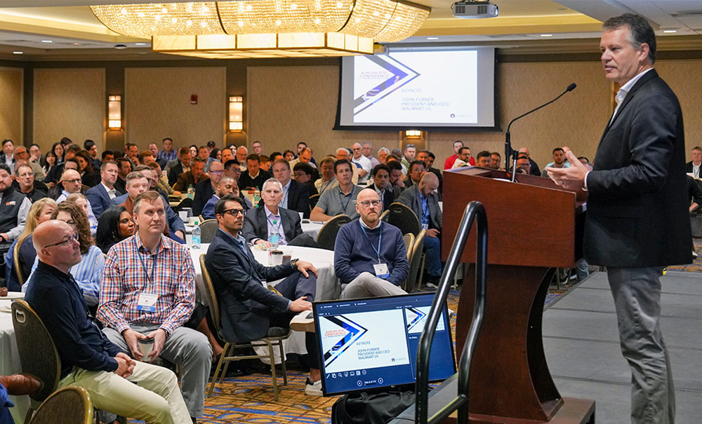 John Furner speaks from a wooden podium to a seated audience in a conference room. A projection screen behind him displays “KEYNOTE JOHN FURNER PRESIDENT AND CEO WALMART US.”