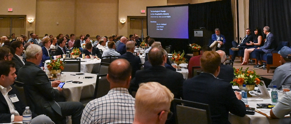 Four panelists sit on a stage during a symposium. A screen beside them reads “Technological Change in the Hospitality Industry.” Attendees sit at round tables in the foreground.
