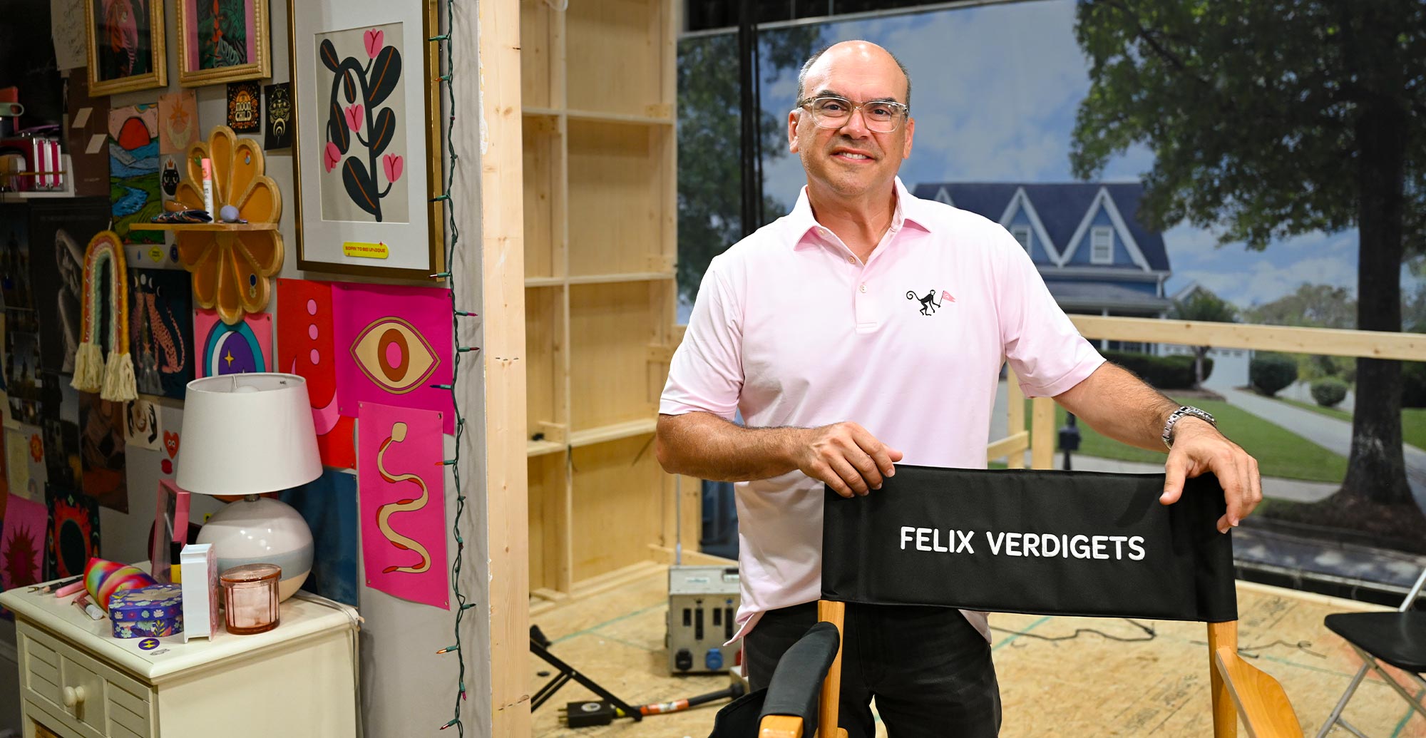 Felix Verdigets stands on a movie set, leaning on a black director’s chair labeled with his name. Behind him is a wall of colorful posters and a scenic backdrop of a suburban house.