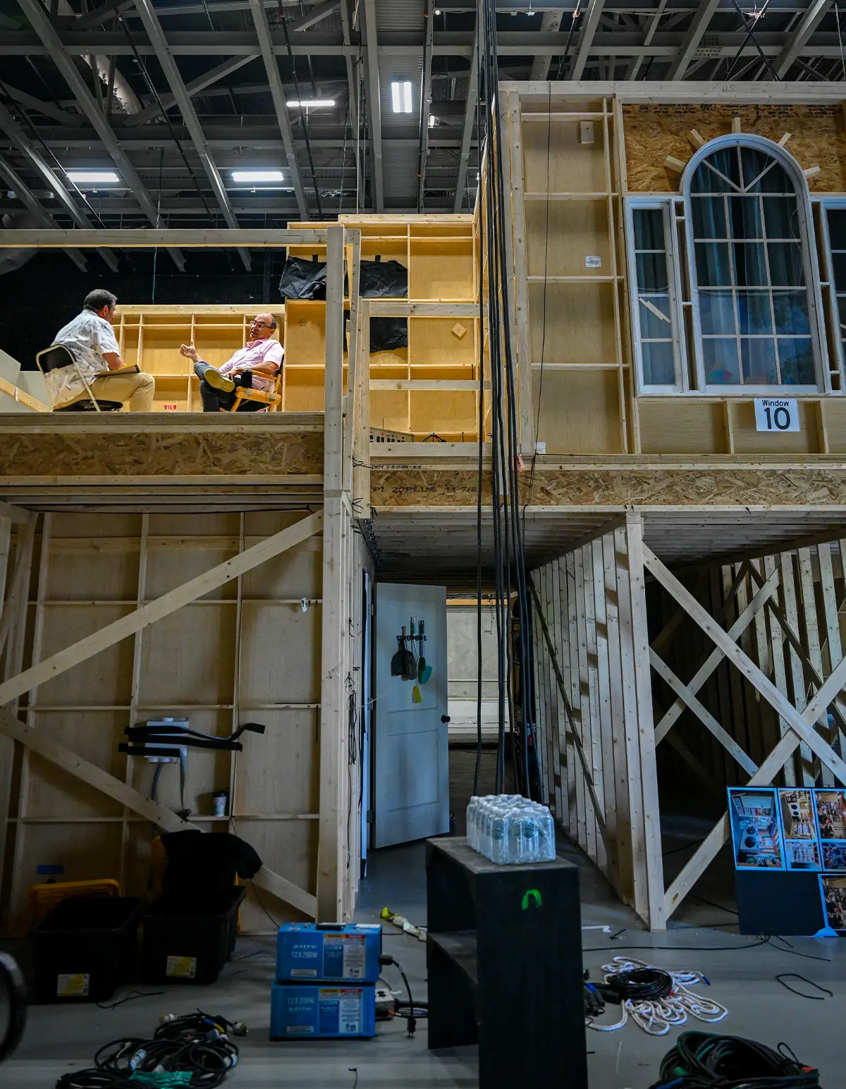 A wide view of a multi-level wooden movie set frame inside a large studio. Two men sit talking on an upper platform, surrounded by exposed timber, hanging cables, and production equipment on the floor below.