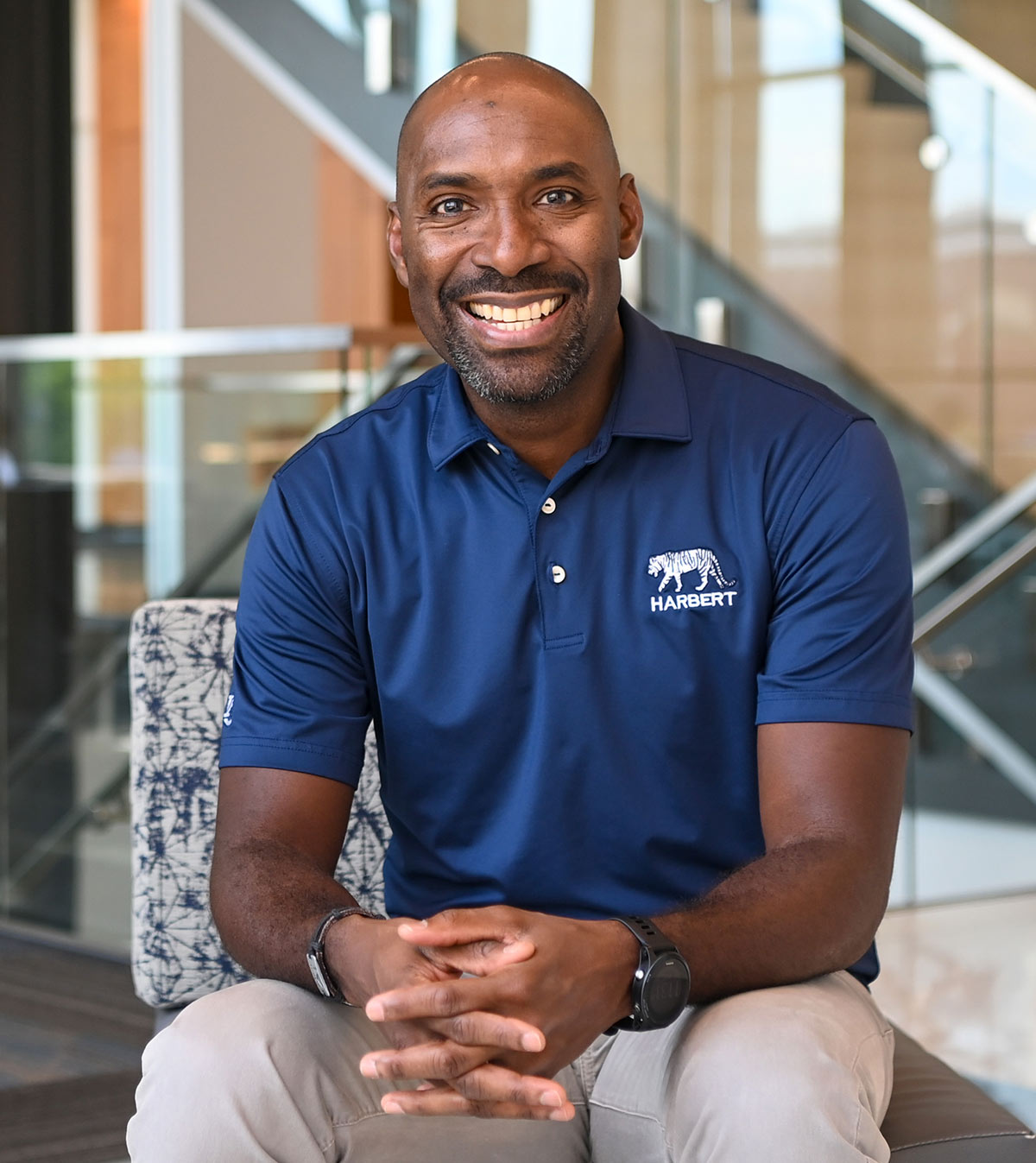 Damion McIntosh wearing a navy polo shirt with a white tiger logo and "HARBERT" text, and khaki pants. An indoor staircase with glass railings is visible in the background.
