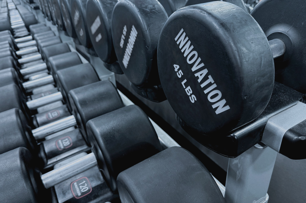 Rows of heavy dumbbells on gym racks, featuring a prominent 45-pound black dumbbell labeled "INNOVATION."