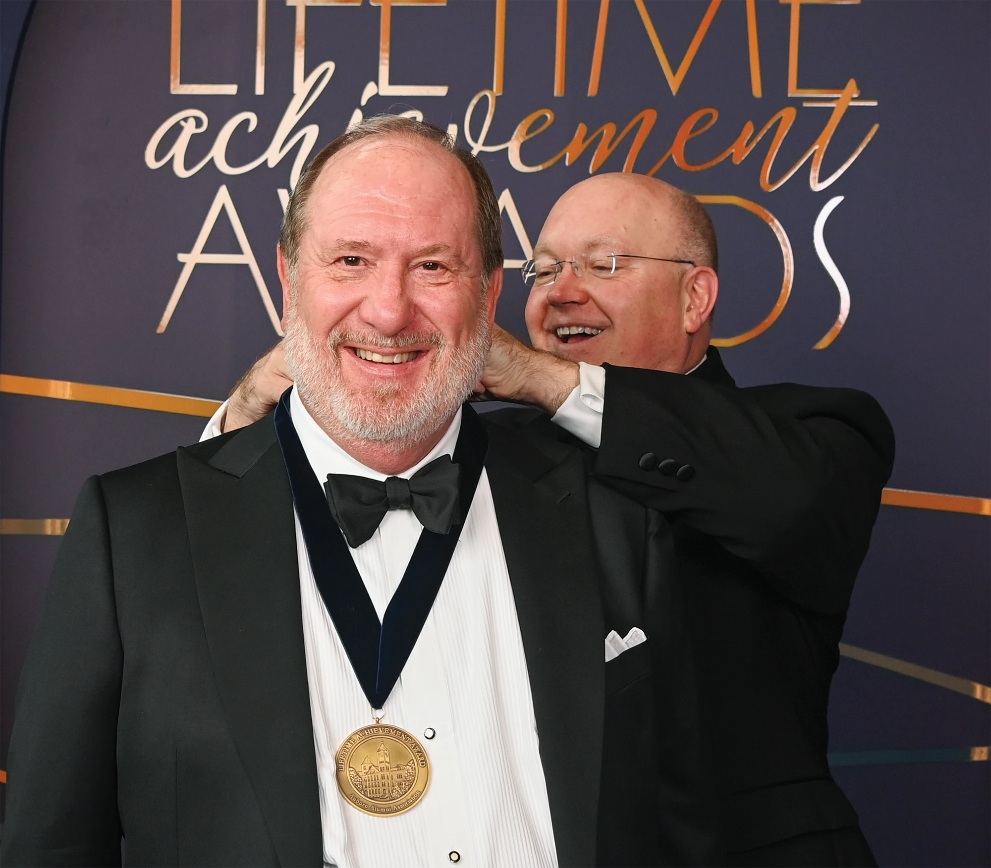Two men in tuxedos smile at an achievement awards event as one wears a medal.