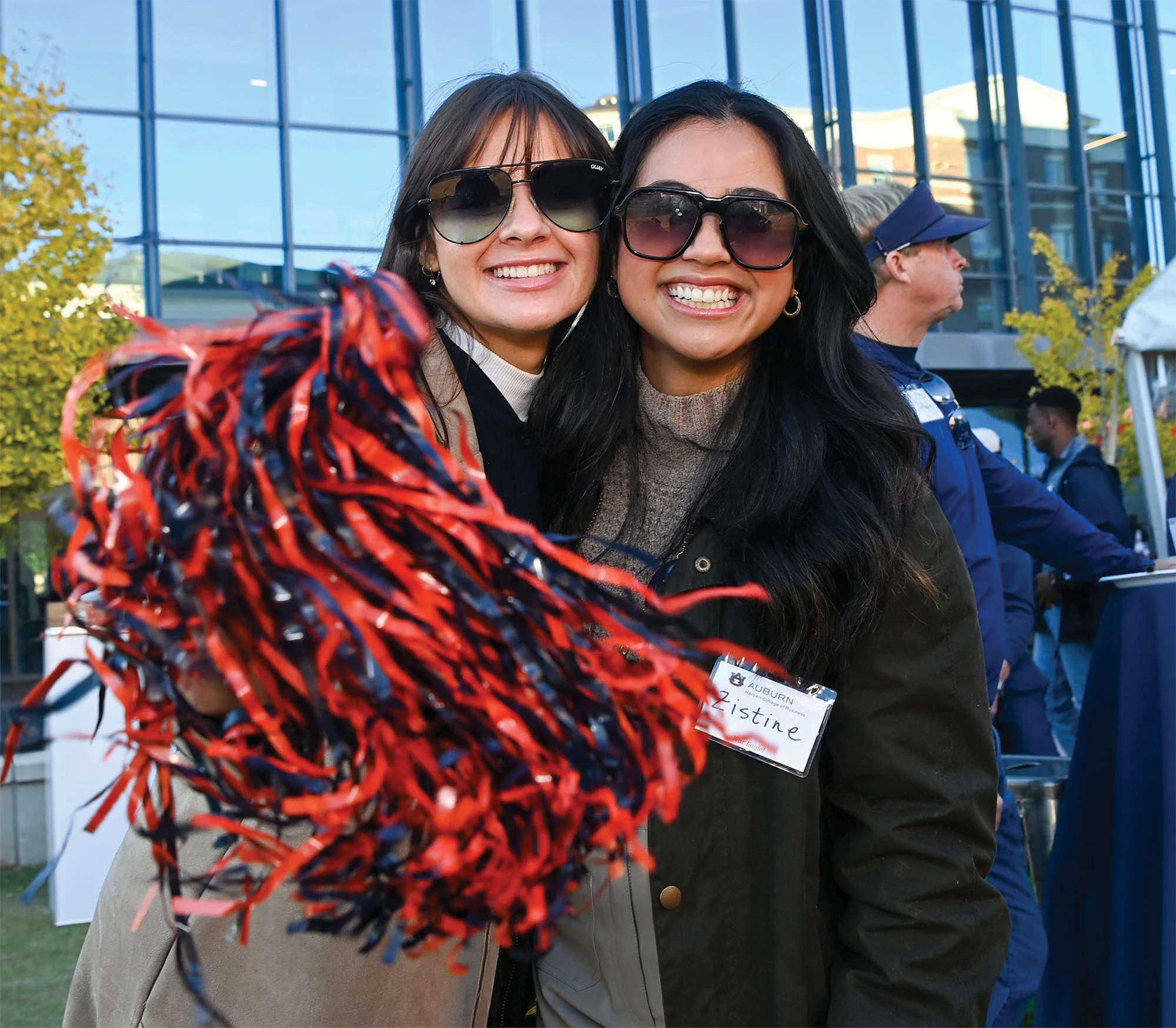 Two women smile while holding Auburn orange and blue pom-poms at an outdoor event.